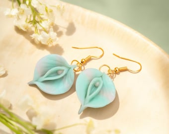 May include: A pair of dangle earrings featuring pale mint green and white calla lily flower shapes made of a smooth material. Each earring is attached to a gold-toned metal hook wire, creating a delicate floral design for ear ornamentation.