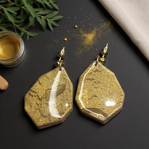 Puede incluir: Un par de pendientes dorados de forma irregular con una textura brillante. Los pendientes tienen cierre de poste y se exhiben sobre una superficie oscura. Un pequeño tarro de purpurina dorada es visible en el fondo.
