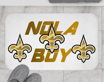 May include: White bath mat with the words "NOLA BOY" in gold lettering. Three gold fleur-de-lis symbols are on the mat.