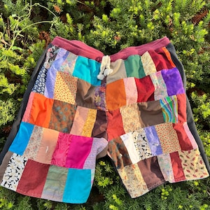 May include: Patchwork shorts with a drawstring waist. The shorts feature a variety of colorful fabric patches in shades of red, orange, blue, and brown. The waistband is burgundy, and the sides are black.