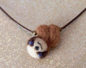 May include: A brown needle-felted sloth pendant on a thin dark brown cord necklace. The sloth figure features a cream-coloured face with dark brown eye patches, small black eyes, and a stitched nose, suspended from a simple brown cord.