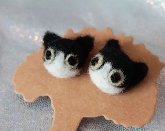 May include: A pair of needle-felted cat head pins featuring black and white wool fibers with prominent light-colored eyes. The two cat heads are attached to a brown, tree-shaped cardboard display card.