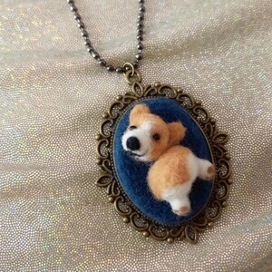 May include: An oval pendant necklace featuring a needle-felted corgi dog figure on a dark blue velvet background, encased in an ornate antique bronze-toned metal frame, suspended from a matching dark metal ball chain necklace.