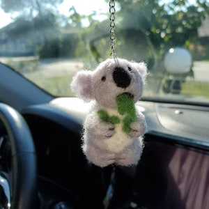 May include: A plush grey koala holding a green leaf, hanging from a rear view mirror in a car.