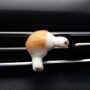 May include: A needle-felted animal rear end ornament featuring white and tan wool fibres. The object has two small legs extending outwards, each tipped with brown circular paw pads, designed to attach to a horizontal vent slat.