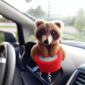 Könnte beinhalten: Ein braunes und weißes Waschbär-Plüschtier hängt an einem Rückspiegel in einem Auto. Der Waschbär sitzt in einem roten und weißen Eimer.