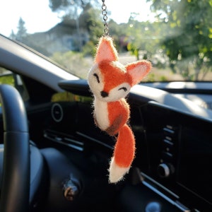 May include: A felt fox car charm hanging from the rearview mirror of a car. The fox is orange and white with a brown nose and black eyes. It is smiling and has a long tail.
