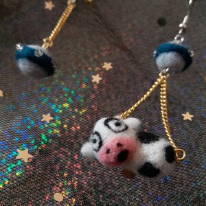 May include: A pair of dangle earrings featuring a black and white felt cow charm and a blue felt sphere with a gold chain.