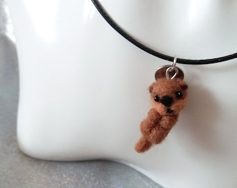 May include: A brown needle-felted otter pendant hangs from a thin black cord. The small, fuzzy otter figure features tiny black bead eyes and a black nose, suspended by a silver-toned metal jump ring attached to the cord.