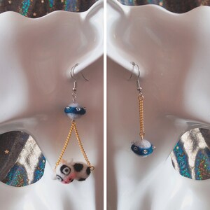 May include: A pair of dangle earrings with a blue and white felt ball charm and a gold chain. The charm has a silver ring around it with rhinestones.