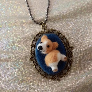 May include: A necklace featuring an oval bronze-toned filigree pendant with a dark blue fabric backing. A small, three-dimensional needle-felted tan and white dog figure is attached to the centre of the pendant, suspended by a metal ball chain.