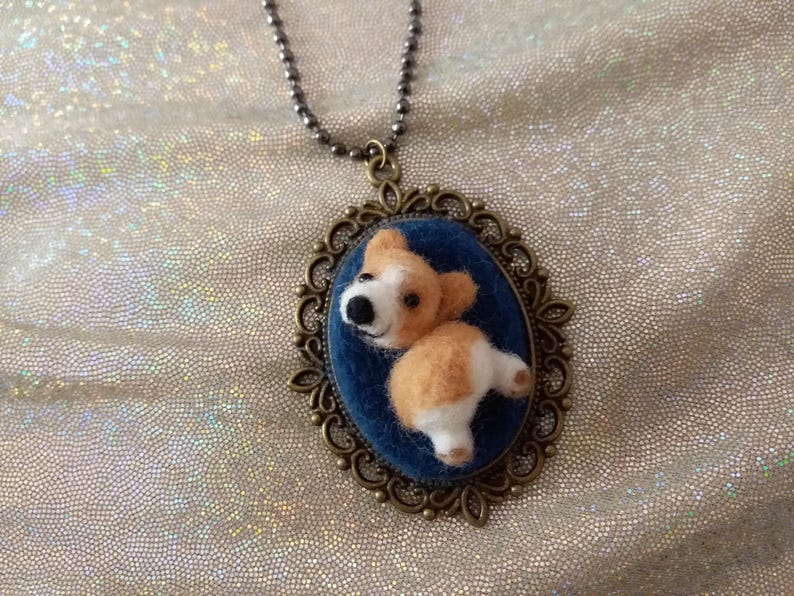 May include: A necklace featuring an oval bronze-toned filigree pendant with a dark blue fabric backing. A small, three-dimensional needle-felted tan and white dog figure is attached to the centre of the pendant, suspended by a metal ball chain.