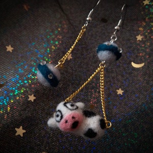 May include: A pair of dangle earrings with a cow charm and a blue and white striped sphere charm. The earrings have gold chains and silver hooks.