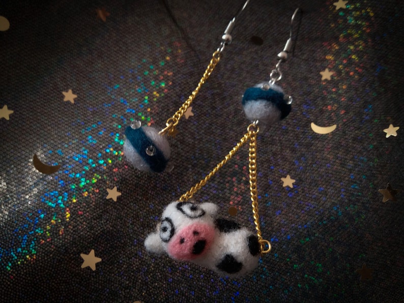 May include: A pair of dangle earrings with a cow charm and a blue and white striped sphere charm. The earrings have gold chains and silver hooks.
