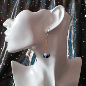 May include: A pair of dangle earrings with a blue and white bead on a gold chain. The earrings are on a white mannequin head.