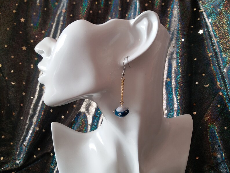 May include: A pair of dangle earrings with a blue and white bead on a gold chain. The earrings are on a white mannequin head.