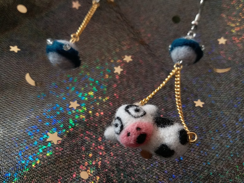 May include: A pair of dangle earrings featuring a black and white felt cow charm and a blue felt sphere with a gold chain.