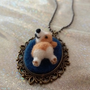 May include: A needle-felted corgi figure with tan and white fur is attached to a dark blue oval felt base. The figure is set within an ornate bronze-toned metal frame pendant that hangs from a matching metallic ball chain necklace.
