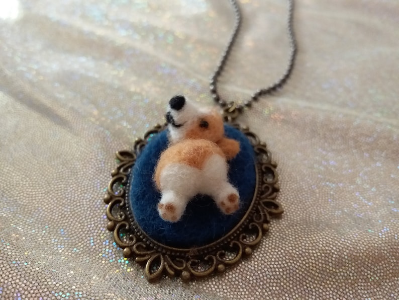 May include: A needle-felted corgi figure with tan and white fur is attached to a dark blue oval felt base. The figure is set within an ornate bronze-toned metal frame pendant that hangs from a matching metallic ball chain necklace.