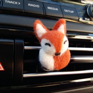 May include: A small orange and white felt fox car air freshener hanging from a car's air vent.