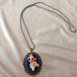 May include: A necklace featuring a bronze-toned oval pendant with an ornate filigree frame. The pendant contains a blue fabric inlay with a small, three-dimensional needle-felted tan and white dog figure attached to a long, thin metal ball chain.