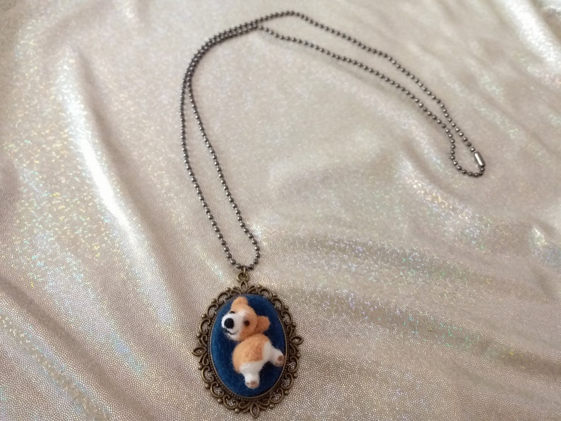 May include: A necklace featuring a bronze-toned oval pendant with an ornate filigree frame. The pendant contains a blue fabric inlay with a small, three-dimensional needle-felted tan and white dog figure attached to a long, thin metal ball chain.