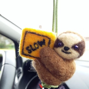 May include: A brown and tan sloth with purple eyes holding a yellow sign that says "SLOW" in black letters. The sloth is hanging from a green string.