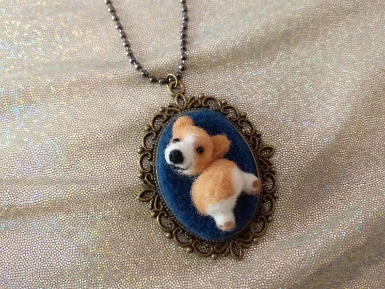May include: An oval pendant necklace featuring a needle-felted corgi dog figure on a dark blue velvet background, encased in an ornate antique bronze-toned metal frame, suspended from a matching dark metal ball chain necklace.