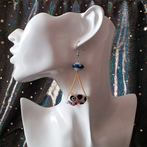 May include: A pair of dangle earrings with a black and white cow print charm hanging from a gold chain. The charm is attached to a blue and white bead.