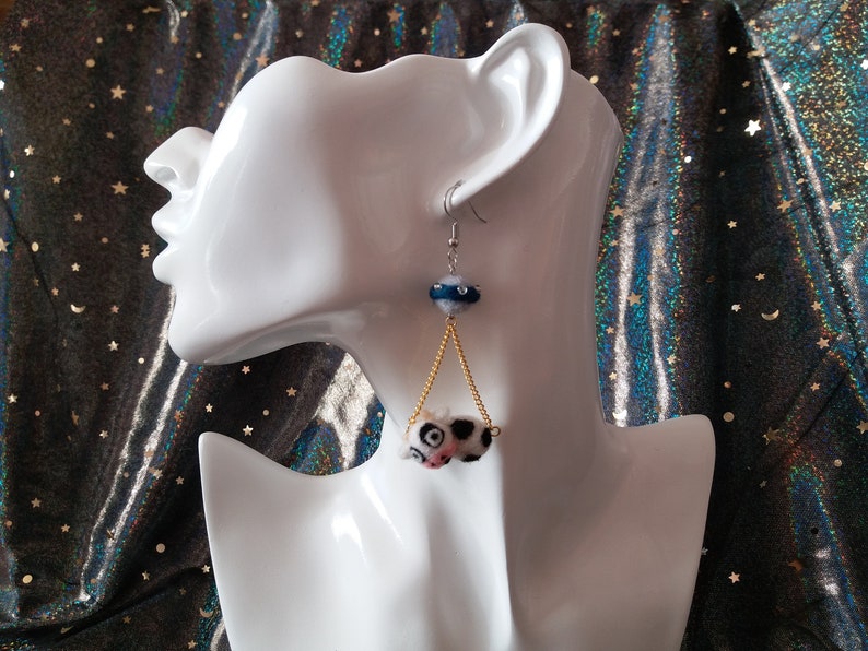 May include: A pair of dangle earrings with a black and white cow print charm hanging from a gold chain. The charm is attached to a blue and white bead.