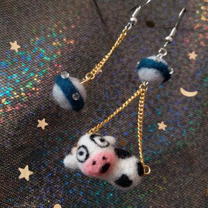 May include: A pair of dangle earrings with a cow charm and a blue and white striped sphere charm. The earrings are made with gold and silver chains.