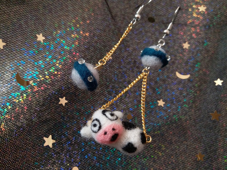May include: A pair of dangle earrings with a cow charm and a blue and white striped sphere charm. The earrings are made with gold and silver chains.