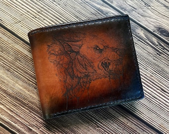 May include: A brown leather bifold wallet featuring a detailed, dark-toned laser-engraved illustration of a snarling wolf head with bared teeth and textured fur, finished with darkened edges and visible stitching along the perimeter.