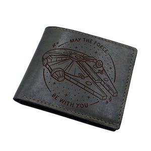 Puede incluir: Una cartera de cuero gris con un diseño grabado con láser marrón del Halcón Milenario, la nave espacial de Star Wars. El texto "Que la fuerza te acompañe" también está grabado en la cartera.