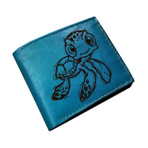 May include: A turquoise leather wallet featuring a cartoon turtle illustration. The wallet is rectangular with a bi-fold design and visible stitching around the edges. The turtle design is etched in dark brown.
