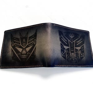 May include: A dark blue and black bi-fold leather wallet featuring two distinct laser-engraved skull-like mask designs on the front panels. The wallet has visible stitching along the edges and a smooth, gradient-toned finish across the surface.