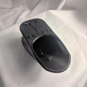 May include: Black 3D-printed Tesla key fob holder. The holder is shaped to cradle the key fob and features the Tesla logo and brand name embossed on the top. The holder is designed to be placed on a flat surface.