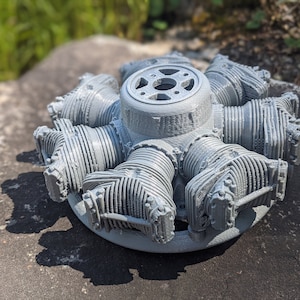 Peut inclure: Un modèle de moteur radial gris imprimé en 3D. Le moteur comporte plusieurs cylindres disposés en cercle autour d'un moyeu central. Le modèle est détaillé, montrant la conception complexe des composants du moteur. Le modèle est posé sur une surface en pierre grise.