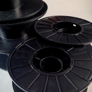 Puede incluir: Primer plano de varias bobinas de plástico negro apiladas y dispuestas. Las bobinas tienen un diseño circular con radios radiales y una abertura central. Estas bobinas probablemente se utilizan para almacenar filamento o alambre.