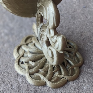 May include: A detailed, gold-colored decorative bracket with an ornate, swirling design. The bracket features a circular base with a floral pattern and a curved support. The overall aesthetic is reminiscent of vintage or antique decor.