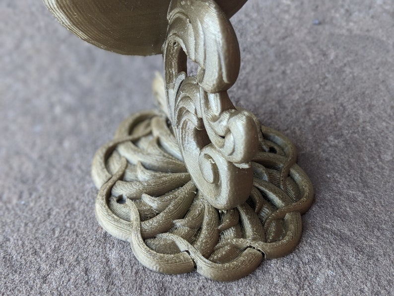 May include: A detailed, gold-colored decorative bracket with an ornate, swirling design. The bracket features a circular base with a floral pattern and a curved support. The overall aesthetic is reminiscent of vintage or antique decor.