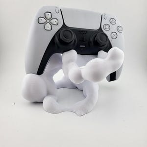 May include: A white and black PlayStation 5 controller resting on a white, abstract-shaped stand. The controller has a symmetrical design with black joysticks and buttons. The stand appears to be 3D-printed.