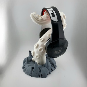 May include: A black and white headset rests on a white tentacle-shaped stand with a gray base. The headset has the word "CU" printed on the headband. The stand is designed to hold the headset.