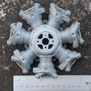 May include: A grey radial engine model with seven cylinders, a central hub, and a ruler for scale. The engine is made of a light grey material and has detailed features. The ruler shows measurements in inches.