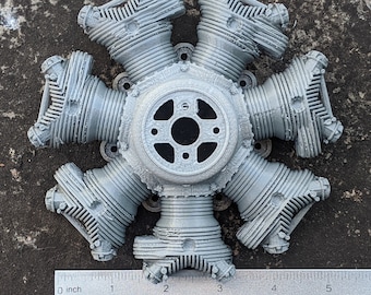 Puede incluir: Un modelo de motor radial gris con siete cilindros, un cubo central y una regla para la escala. El motor está hecho de un material gris claro y tiene detalles. La regla muestra medidas en centímetros.