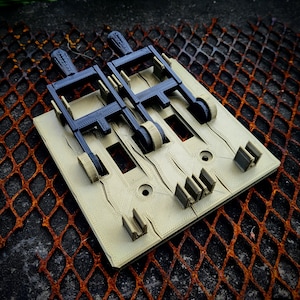 以下が含まれることがあります： 2つのレバーのような機構といくつかの長方形のコンポーネントを備えた、ベージュと黒の3Dプリントオブジェクト。オブジェクトは、錆びた金属グリッドの背景を持つテクスチャ加工された表面に配置されています。