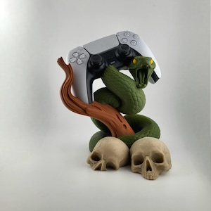 Puede incluir: Un mando de videojuegos blanco y negro descansa sobre una escultura de serpiente verde con ojos amarillos, enrollada alrededor de una rama marrón. Dos calaveras beige en la base, un accesorio de juego único.