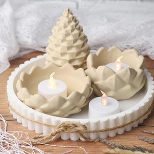 Puede incluir: Una bandeja decorativa blanca con borde festoneado sostiene tres candelabros beige y un adorno geométrico en forma de árbol. Cada candelabro contiene una pequeña vela a pilas con una llama parpadeante, creando un ambiente cálido.