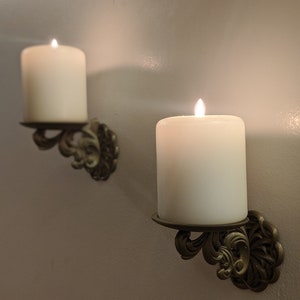 May include: Two ivory-colored pillar candles with flickering flames are mounted on ornate, antique-style wall sconces. The sconces are made of a bronze-colored metal and feature a decorative, scrolled design. The candles are set against a neutral-colored wall.