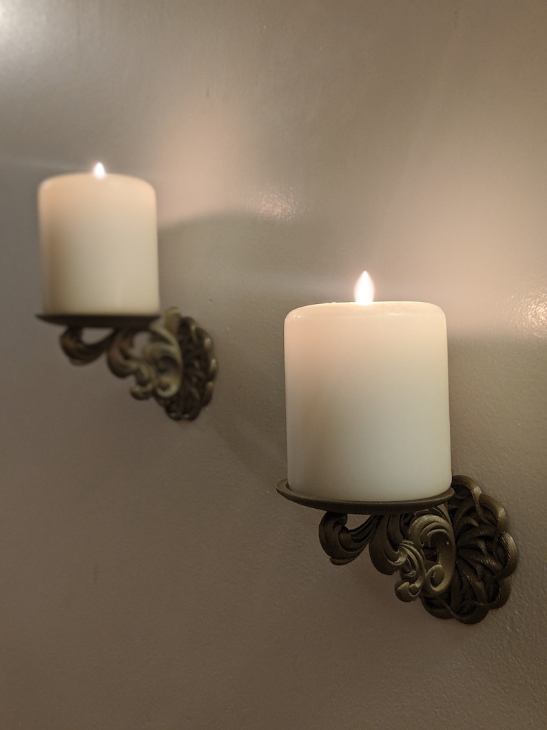 May include: Two ivory-colored pillar candles with flickering flames are mounted on ornate, antique-style wall sconces. The sconces are made of a bronze-colored metal and feature a decorative, scrolled design. The candles are set against a neutral-colored wall.